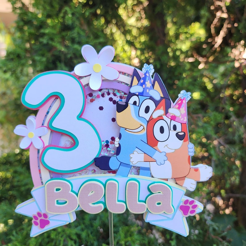 May include: A vibrant cake topper with the number "3" and the name "Bella". It features cartoon characters, a rainbow, flowers, and confetti. The characters are embracing. The topper is mounted on a stick.