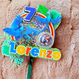 Op de afbeelding: Een kleurrijke taarttopper met een blauwe ara, een regenboog en het getal 7. De naam "Lorenzo" staat in feloranje en gele letters. De topper heeft een groene steel om in een taart te steken.