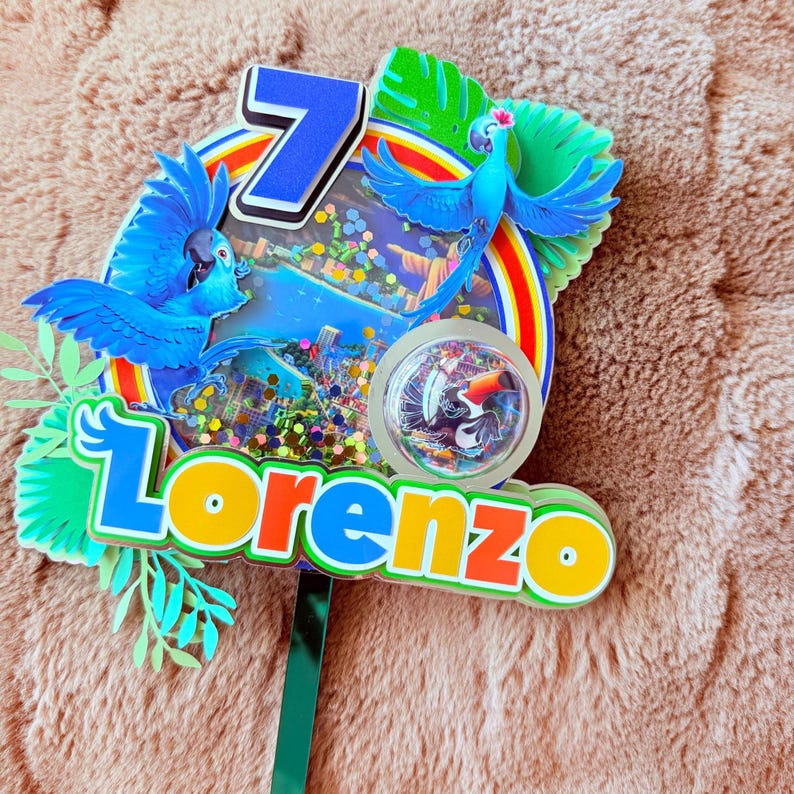 Op de afbeelding: Een kleurrijke taarttopper met een blauwe ara, een regenboog en het getal 7. De naam "Lorenzo" staat in feloranje en gele letters. De topper heeft een groene steel om in een taart te steken.