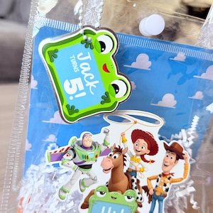 May include: A clear plastic favor bag filled with white paper shred, featuring a blue sky and cloud background. Attached are cutouts of Buzz Lightyear, Jessie, Woody, and Bullseye, along with two green frog signs reading Jack turns 5! and Hi! Let's play!