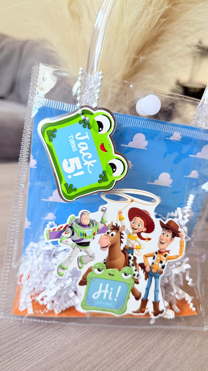 May include: A clear plastic favor bag filled with white paper shred, featuring a blue sky and cloud background. Attached are cutouts of Buzz Lightyear, Jessie, Woody, and Bullseye, along with two green frog signs reading Jack turns 5! and Hi! Let's play!