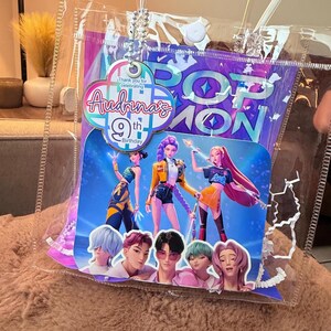 Puede incluir: Una bolsa de regalo de pl&aacute;stico transparente con un tema de Pop Icon. La bolsa presenta un gr&aacute;fico colorido de personajes animados y el texto "9&ordm; cumplea&ntilde;os de Audrina". Una cadena plateada y un bot&oacute;n blanco est&aacute;n unidos a la bolsa.