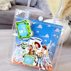 May include: A clear plastic gift bag with a white snap closure and clear handles. Inside is a blue background with white clouds, white paper shred, and character cutouts of Buzz Lightyear, Jessie, Woody, and Bullseye. Tags read Jack Turns 5! and Hi!