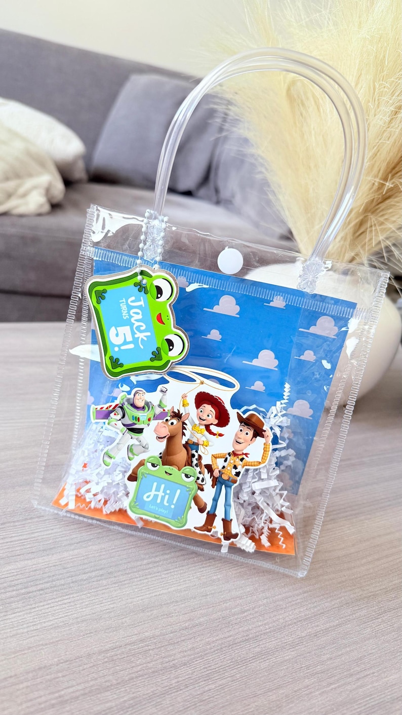 May include: A clear plastic gift bag with a white snap closure and clear handles. Inside is a blue background with white clouds, white paper shred, and character cutouts of Buzz Lightyear, Jessie, Woody, and Bullseye. Tags read Jack Turns 5! and Hi!