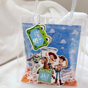 May include: A clear plastic gift bag with two transparent handles and a snap closure. The bag features a blue sky and cloud background with Toy Story character cutouts, including Woody, Jessie, Bullseye, and Buzz Lightyear. A green frog tag reads Jack turns 5! and Hi! Let's play!