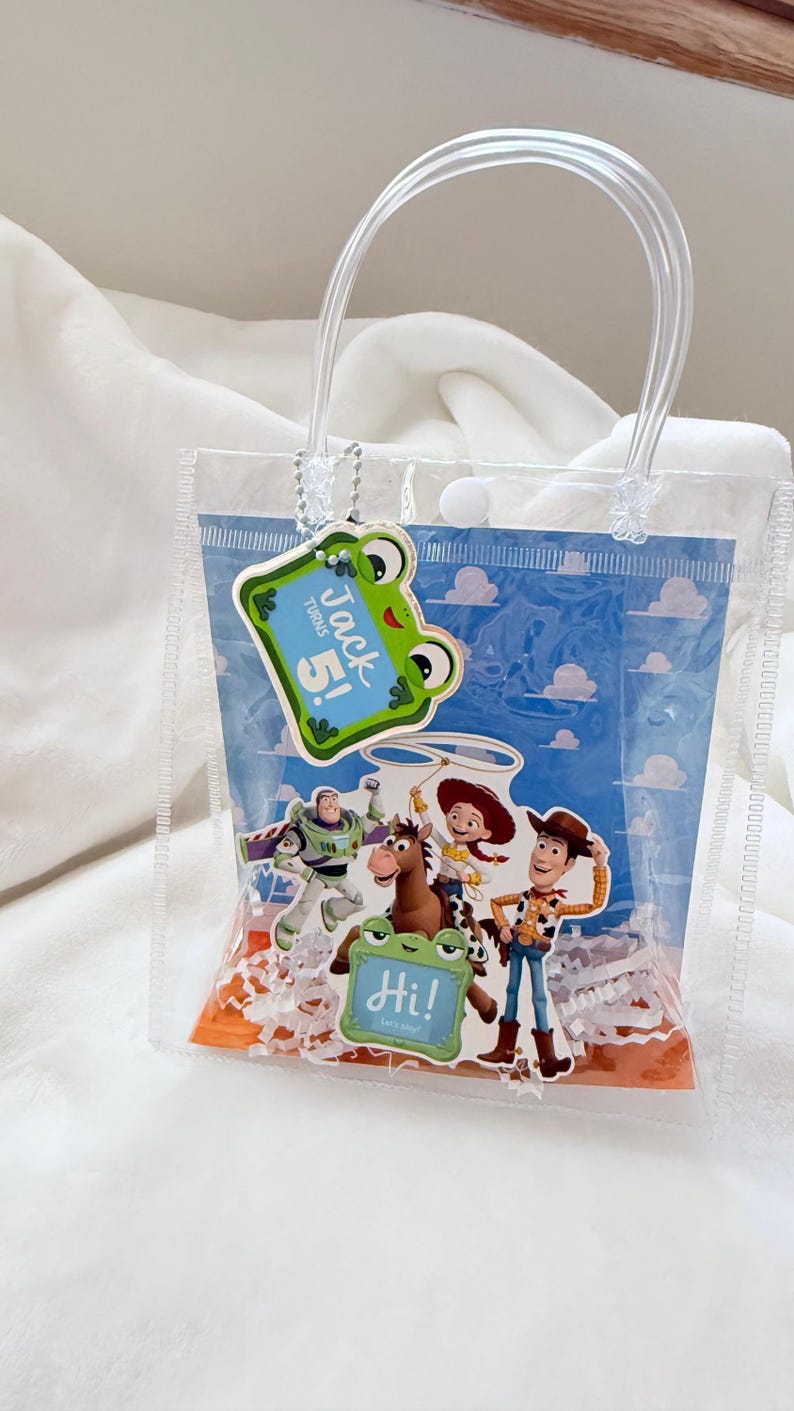 May include: A clear plastic gift bag with two transparent handles and a snap closure. The bag features a blue sky and cloud background with Toy Story character cutouts, including Woody, Jessie, Bullseye, and Buzz Lightyear. A green frog tag reads Jack turns 5! and Hi! Let's play!