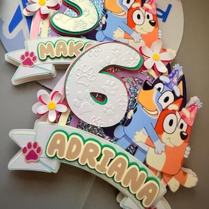 May include: A vibrant birthday cake topper with the number 6, the name "Adriana", and cartoon characters. The design incorporates paw prints, flowers, and the word "MAK". The characters are blue and orange.