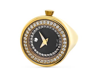 May include: A gold ring with a black watch face surrounded by a ring of clear rhinestones. The watch face has a white hour and minute hand and a small white dot at the 12 o'clock position. The watch face also has the word "MOJID" printed on it.