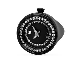 Pode incluir: Um relógio de pulso preto com uma moldura encrustada com diamantes. O mostrador do relógio é preto com ponteiros prateados e um diamante prateado na posição das 12 horas. O mostrador do relógio também tem a palavra "MOJID" impressa.
