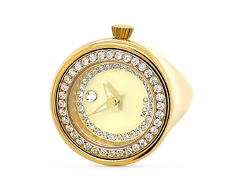 Pode incluir: Um relógio de anel de ouro com um mostrador redondo e uma moldura encrustada com diamantes. O mostrador do relógio é branco com ponteiros pretos e um pequeno diamante na posição das 12 horas. O mostrador do relógio também tem a palavra "MOUJD" impressa nele.