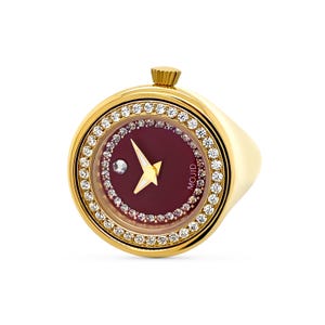 May include: A gold ring with a round watch face set with clear rhinestones. The watch face has a burgundy background with a white and gold arrow-shaped hand and a small diamond at the centre. The watch face also has the word "MOJID" printed on it.