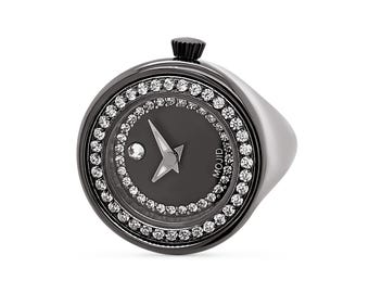 Pode incluir: Um relógio de anel preto com um mostrador redondo e uma moldura encrustada com diamantes. O mostrador do relógio tem um fundo preto com ponteiros brancos e uma estrela branca. O mostrador do relógio também tem a palavra "MOJID" impressa nele.