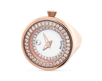 Pode incluir: Um relógio de anel de ouro rosa com um mostrador branco e um círculo de cristais transparentes ao redor da borda. O mostrador do relógio tem um pequeno diamante na posição das 12 horas e a palavra "MOJID" impressa no lado direito.