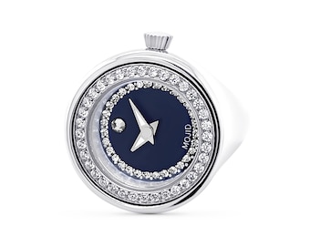 Pode incluir: Um anel de prata com um relógio de mostrador azul. O mostrador do relógio tem uma estrela prateada e um único diamante. O anel é decorado com um círculo de pequenos diamantes.