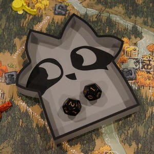 May include: A gray raccoon-shaped dice tray with black accents and two black dice with gold numbers. The tray sits on a game board with a forest and village scene, with orange and yellow game pieces.
