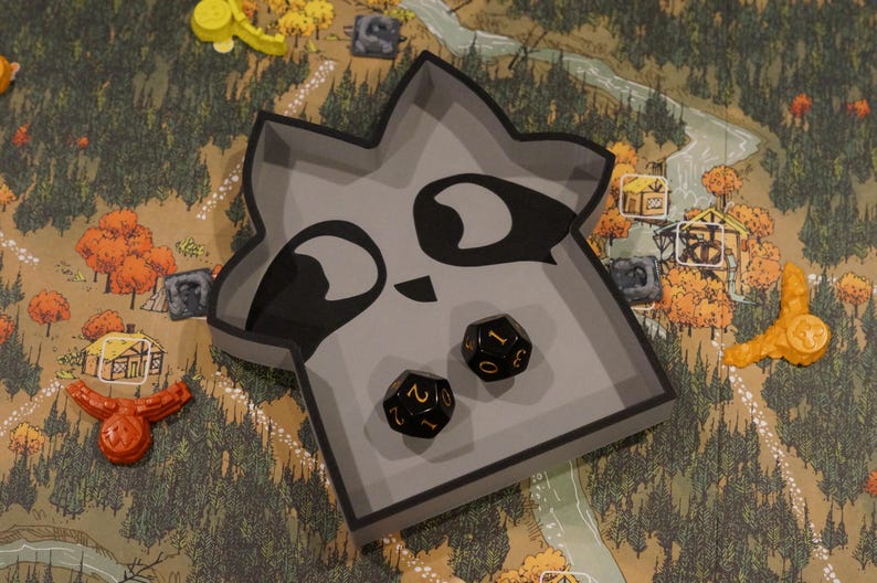 May include: A gray raccoon-shaped dice tray with black accents and two black dice with gold numbers. The tray sits on a game board with a forest and village scene, with orange and yellow game pieces.