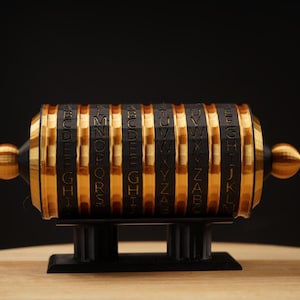May include: A black and gold cipher wheel with rotating lettered bands. The bands are gold with black lettering and black vertical stripes. The cipher wheel sits on a black stand with a wooden surface in the foreground and a black background.