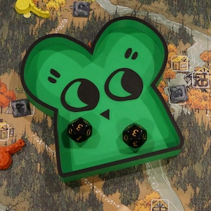 May include: A green, cartoon-faced dice tray with two black dice inside, set on a game board depicting a forest scene. The tray has a black outline and a whimsical design. Other game pieces are scattered around.