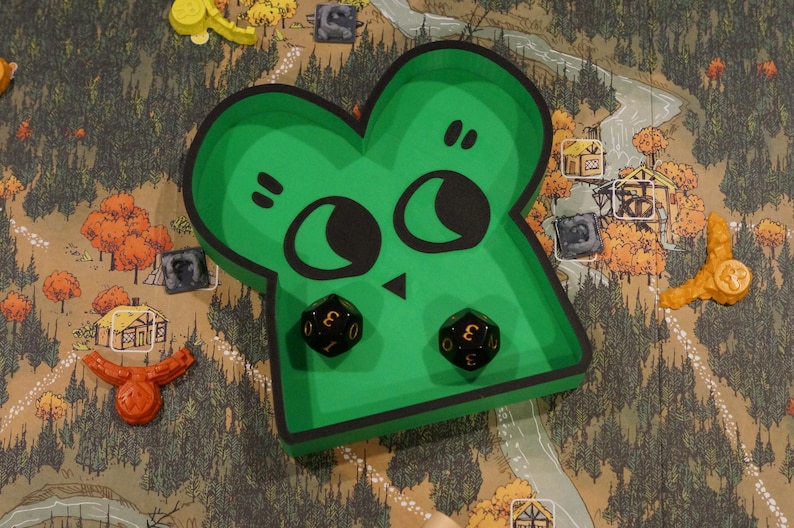 May include: A green, cartoon-faced dice tray with two black dice inside, set on a game board depicting a forest scene. The tray has a black outline and a whimsical design. Other game pieces are scattered around.