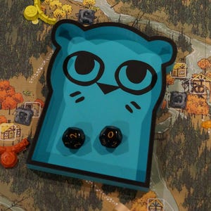 May include: A teal-colored game piece shaped like an owl with black eyes and whiskers, holding two black dice with white numbers. The piece sits on a game board with a forest and village design, along with other game pieces.