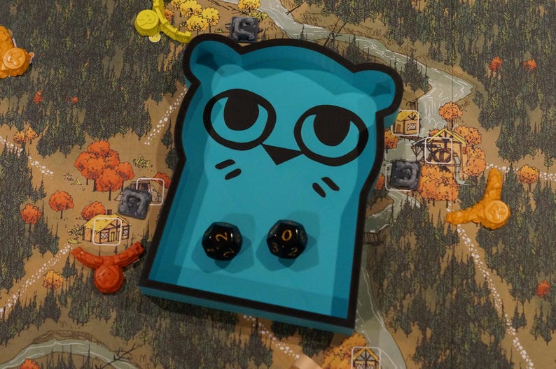 May include: A teal-colored game piece shaped like an owl with black eyes and whiskers, holding two black dice with white numbers. The piece sits on a game board with a forest and village design, along with other game pieces.