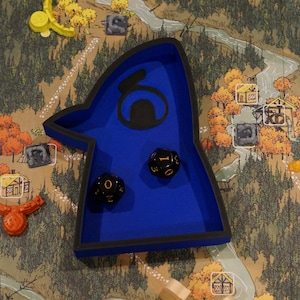 May include: A blue dice tray with a black border and a stylized design, holding two black dice with white numbering. The tray sits on a game board depicting a map with trees, a river, and game pieces.