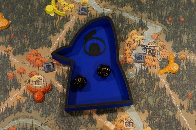 May include: A blue dice tray with a black border and a stylized design, holding two black dice with white numbering. The tray sits on a game board depicting a map with trees, a river, and game pieces.