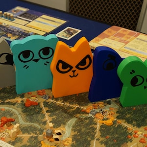 May include: A board game with colorful wooden animal figures. The game pieces are shaped like cats, a raccoon, and a frog, with black facial features. The game board features a map with various terrain elements. Other game components are visible.