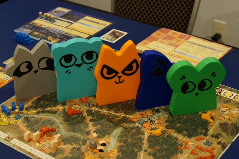 May include: A board game with colorful wooden animal figures. The game pieces are shaped like cats, a raccoon, and a frog, with black facial features. The game board features a map with various terrain elements. Other game components are visible.