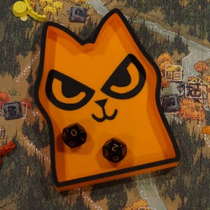 May include: Orange cat-shaped dice tray with black accents, holding two black dice. The tray sits on a game board depicting a forest scene with houses and tokens. A fun accessory for tabletop gaming.
