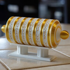 May include: A white and gold cipher wheel with rotating lettered bands. The bands are gold with white lettering and are mounted on a white stand. The ends of the wheel are gold spheres. The cipher wheel is a puzzle or decorative item.