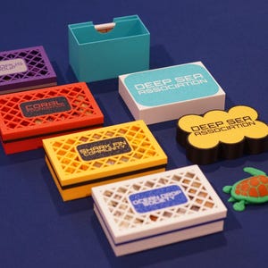 May include: A selection of small, rectangular boxes in various colours, featuring lattice tops and labels for ocean-themed groups. A teal box and a yellow, cloud-shaped item are also visible, alongside a small turtle figurine.