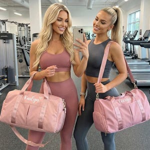 Könnte beinhalten: Zwei Frauen in einem Fitnessstudio, jede mit einer rosa Sporttasche. Die Taschen haben einen Schultergurt und die Namen "Kailyn" und "Georgia" sind aufgedruckt. Die Frauen tragen Trainingskleidung in Rosa- und Grautönen.
