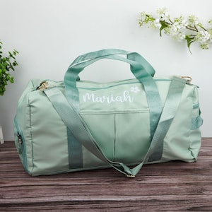Könnte beinhalten: Eine hell salbeigrüne Reisetasche mit dem Namen Mariah in weißer Schreibschrift und einem kleinen weißen Blumen-Symbol. Die Tasche verfügt über zwei Tragegriffe, einen abnehmbaren Schultergurt und eine Vordertasche mit glatter Oberfläche.