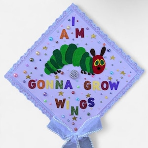 May include: A graduation cap decorated with a green and red caterpillar design. The cap has the words "I AM GONNA GROW WINGS" in colourful letters, with star and bead embellishments, and a light blue bow.
