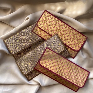 Puede incluir: Varios sobres decorativos con patrones geométricos y de ondas doradas. Los sobres tienen un borde burdeos y están dispuestos sobre una superficie satinada. Los sobres son de varios tonos de marrón y dorado.