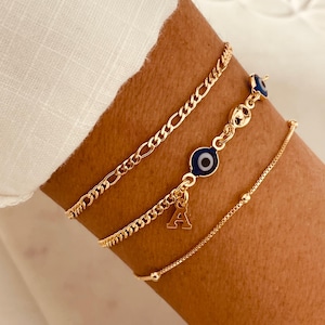 May include: A set of three gold-toned bracelets. The top bracelet is a classic figaro chain. The middle bracelet features a series of small blue and white evil eye charms and a dangling letter A charm. The bottom bracelet is a delicate box chain.