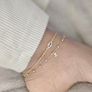 Może przedstawiać: Dwie złote bransoletki na kostkę z niebieskim amuletem w kształcie oka i małym krzyżykiem na jednej bransoletce oraz literą 'N' na drugiej.