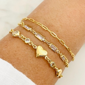 May include: Three gold bracelets with clear crystal accents. One bracelet features a heart charm, another has rectangular crystals, and the third is a simple chain.