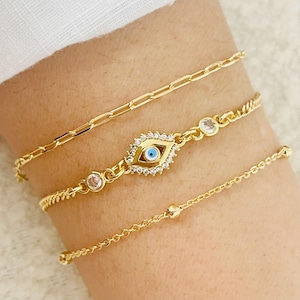 May include: Three gold-colored bracelets. One is a chain link style, another features an evil eye charm with a blue center and clear stone accents, and the third is a delicate chain with small beads. The bracelets are worn on a wrist.