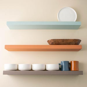 May include: Three shelves with different colored paint. The top shelf is light blue, the middle shelf is orange, and the bottom shelf is gray. The top shelf has a white plate on it. The middle shelf has a wooden bowl on it. The bottom shelf has five white bowls and two mugs, one blue and one orange.
