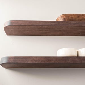 May include: Two dark brown wooden floating shelves mounted on a white wall. The top shelf holds a wooden bowl and a white plate. The bottom shelf displays several white ceramic bowls. Modern home decor.