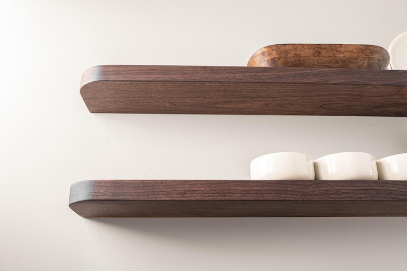 May include: Two dark brown wooden floating shelves mounted on a white wall. The top shelf holds a wooden bowl and a white plate. The bottom shelf displays several white ceramic bowls. Modern home decor.