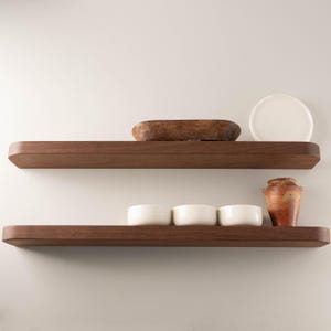 May include: Two dark brown wooden floating shelves with rounded edges. The top shelf holds a wooden bowl and a white plate. The bottom shelf displays three white bowls and a small, textured, orange and brown vase.