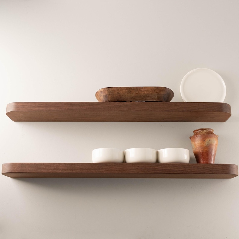 May include: Two dark brown wooden floating shelves with rounded edges. The top shelf holds a wooden bowl and a white plate. The bottom shelf displays three white bowls and a small, textured, orange and brown vase.