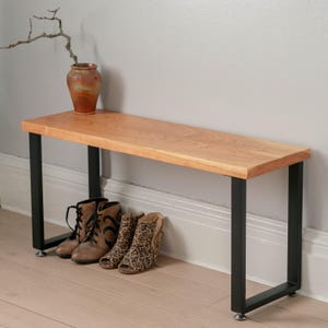 May include: A wooden bench with black metal legs. The bench is made of light wood and has a natural finish. The legs are square and have a black powder coat finish. The bench is sitting on a wooden floor. There are three pairs of shoes under the bench.