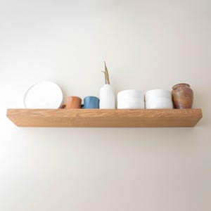 May include: A wooden shelf displays a white plate, two small mugs in brown and blue, a white ribbed vase with a dried stem, two white lidded containers, and a brown ceramic jar. The shelf is made of light-colored wood and is mounted on a white wall.