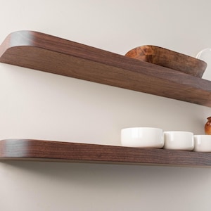 May include: Two dark brown wooden floating shelves mounted on a light-colored wall. The top shelf holds a wooden bowl and a white plate. The bottom shelf displays three white bowls and a brown vase.