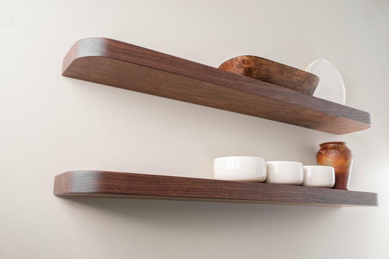 May include: Two dark brown wooden floating shelves mounted on a light-colored wall. The top shelf holds a wooden bowl and a white plate. The bottom shelf displays three white bowls and a brown vase.