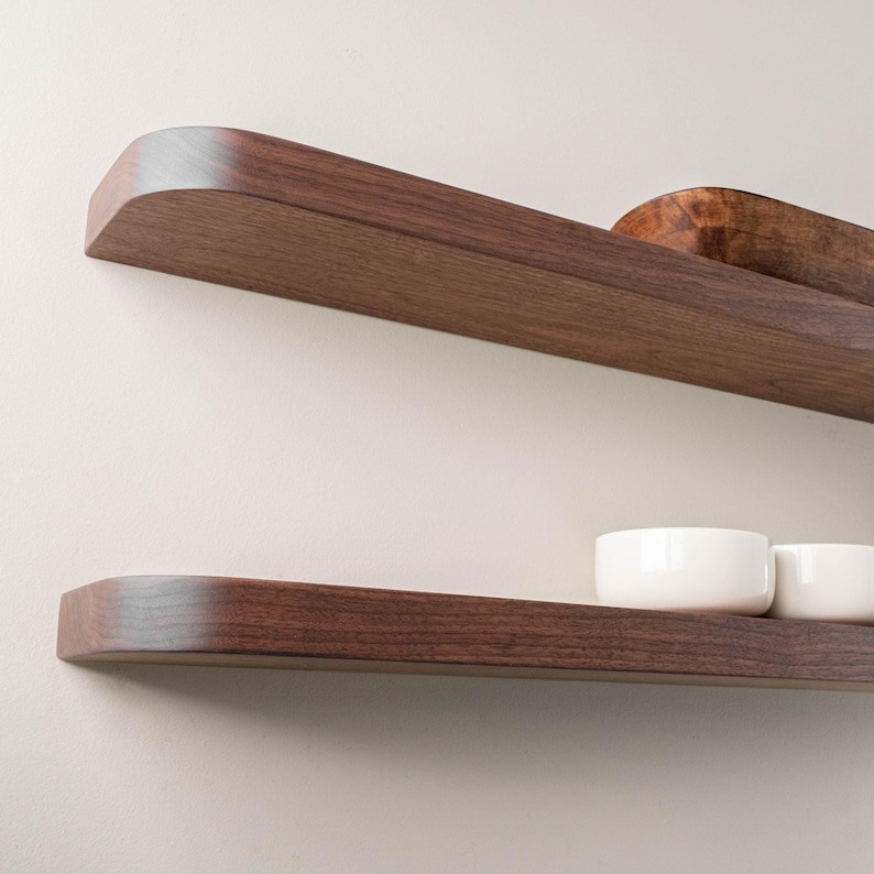 May include: Two floating wooden shelves with a rich, dark brown finish. The top shelf holds a small, wooden bowl. The bottom shelf displays two white ceramic bowls. The shelves have rounded edges and are mounted on a neutral-colored wall.